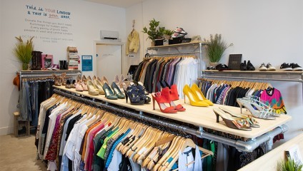 A thrift store interior featuring racks of clothing and a central display of various shoes, including high heels and sandals. A wall quote reads, This is your London and this is your shop. The store is well-lit and organized, with plants decorating the space.