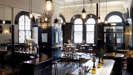 Inside the pub. dark wooden bar, tables and chairs and swing lights. Large victorian windows.