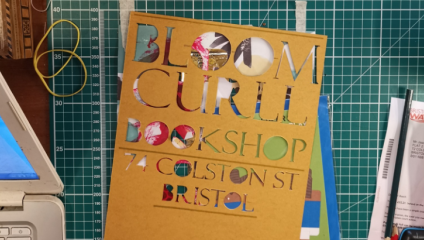 A cardboard template placed on a green cutting mat, with cut-out letters revealing colorful designs underneath. The text reads Bloom Curll Bookshop, 74 Colston St, Bristol. A pen, letter, laptop edge, and other desk items are partially visible.