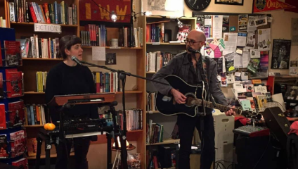 Two musicians perform in a cozy bookstore with shelves of books behind them. The woman on the left plays a keyboard and sings into a microphone, while the man on the right plays an acoustic guitar and sings. The background is filled with posters and books.