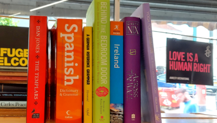 A row of books on a shelf with titles including The Templars, Spanish Dictionary & Grammar,A German Reference Grammar, Behind the Bedroom Door, Ireland, Across That Wall, and Yoga For Wellness. A sign nearby reads, Love is a Human Right.