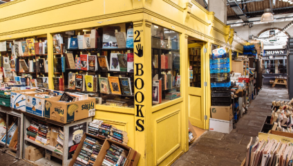 A bustling indoor market showcasing a vibrant yellow stall filled with an array of colorful books. Signs display prices, and stacks of both new and used books line the shelves and tables. The surrounding area is crowded with more book-filled stalls.