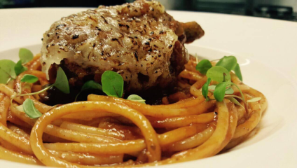 A close-up of a gourmet plate featuring a well-seasoned and grilled piece of meat on top of a bed of sauced spaghetti. The dish is garnished with fresh green microgreens, presented on a white plate.