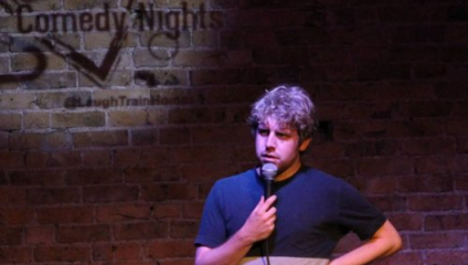 A man with grayish hair performs stand-up comedy on stage, holding a microphone in one hand and resting the other on his hip. The background features a brick wall with projected text that reads Comedy Nights and a Twitter handle @LaughTrainHome.