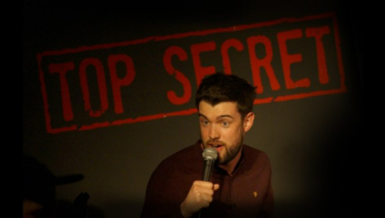 A comedian with a beard and short hair performs on stage, holding a microphone. Behind him is a backdrop with the words TOP SECRET in large, red letters. The background is mostly dark, focusing attention on the performer.