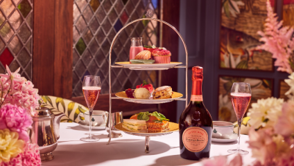 A three-tiered stand displaying a variety of desserts and pastries, surrounded by two glasses of sparkling rosé and a bottle of rosé champagne. The table is adorned with pink and yellow flowers, and there are teacups and a teapot. Stained glass windows in the background.