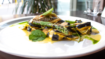 A close-up of an elegantly plated dish on a white plate, featuring ravioli with black and white striped pasta. It is garnished with green asparagus spears, herbs, and a creamy yellow sauce. In the background, a blurred setting includes a glass and potted plant.