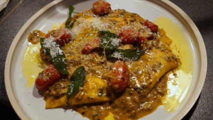 A plate of ravioli covered in a creamy, meat-based sauce with cherry tomatoes, fresh herbs, and grated cheese. The yellow ravioli and sauce are garnished with oil and sage leaves. The dish is served on a white plate with a light brown rim.