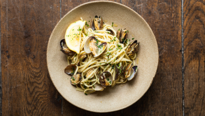 A bowl of spaghetti alle vongole garnished with chopped herbs and a lemon slice, placed on a wooden table. The dish consists of spaghetti noodles mixed with clams in their shells.