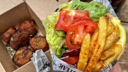 A close-up of a flavorful wrap filled with fresh lettuce, sliced tomatoes, fried potatoes, and various toppings. Next to it is an open box containing crispy, golden-brown fritters garnished with chopped herbs. The meal appears appetizing and ready to eat.