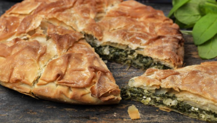 A close-up of a golden-brown spinach pie on a dark wooden surface. One slice has been cut and placed next to the pie, revealing the spinach and cheese filling. Fresh green leaves are seen in the background.