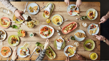 A wooden table is filled with plates of colorful Mediterranean foods, including wraps, salads, hummus, and flatbreads. Multiple hands reach in to serve or eat the dishes, and drinks like wine and beer are also visible on the table.