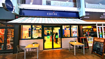 Street view of the facade of Kouzina, a cozy restaurant with a blue sign and white awning. Two tables with chairs are positioned outside, and the restaurant's interior can be seen through the glass doors. Surrounding businesses include Bento Boss and Sushi & Pan.
