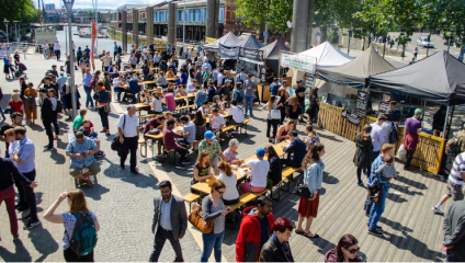A bustling outdoor market next to a waterfront. Numerous people are gathered around various food stalls under black tents, sitting at communal tables, and walking along the wooden boardwalk. Nearby buildings and trees are visible under a sunny blue sky.