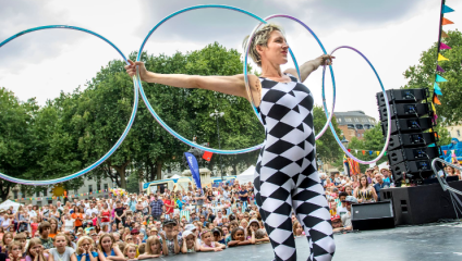 A performer in a black and white checkered bodysuit is balancing several large hula hoops on her arms on an outdoor stage. A large, attentive crowd is watching in the background, surrounded by trees and colorful bunting on a sunny day.