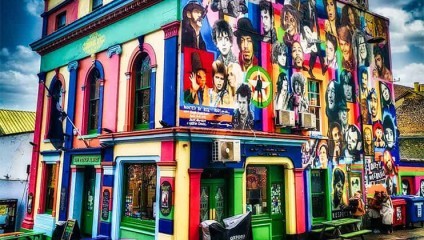 A vibrantly painted building features a colorful mural with numerous portraits of famous musicians and cultural icons. The building, adorned with bright blues, pinks, greens, and oranges, has green-painted windows and doors, standing out against a partly cloudy sky.