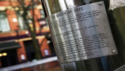 A close-up of a metallic plaque on a monument titled The Beacon of Hope, dedicated to individuals and families affected by HIV/AIDS. The background shows a blurred view of buildings and trees. The plaque lists contributors to the project's completion.