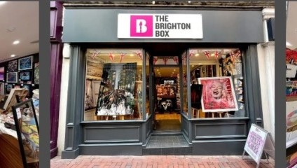 A storefront labeled The Brighton Box is displayed centrally. The interior features various art pieces, framed photographs, and artwork on both walls and center display racks. The store appears well-lit and colorful, showcasing a wide range of creative works.