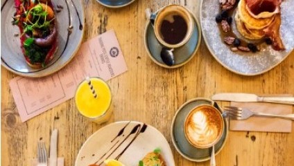 Top-down view of a wooden table set with various breakfast dishes including pancakes with bacon and blueberries, toast topped with smoked salmon and cream cheese, a plate with a variety of sausages and vegetables, and drinks like coffee and a smoothie.
