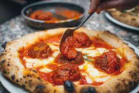 Close-up of a freshly baked pizza with a golden crust, topped with tomato sauce, melted cheese, and large meatballs. A hand holding a spoon is adding more sauce to one of the meatballs. A pan with additional meatballs in sauce is visible in the background.