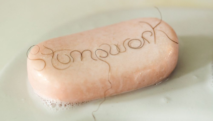 A bar of pink soap with the word stompwork spelled out on its surface using strands of hair. The soap is slightly wet with soap suds visible around its base on a light-colored surface.