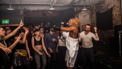 A performer with blonde hair and a white outfit entertains an energetic crowd at a lively indoor venue. Audience members cheer, raise their hands, and engage enthusiastically. The setting is intimate with industrial decor, and the atmosphere is vibrant and dynamic.