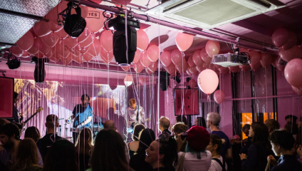 A lively indoor concert features musicians performing on stage beneath a ceiling adorned with numerous pink balloons. Attendees are gathered closely, enjoying the music amidst the festive atmosphere, with vibrant lighting enhancing the scene.