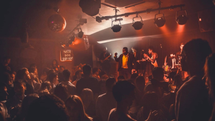A crowded music venue with a live band performing on stage. The band is bathed in warm orange and red lights, creating an intimate atmosphere. People in the audience are dancing and enjoying the music, some holding drinks. A disco ball hangs from the ceiling.