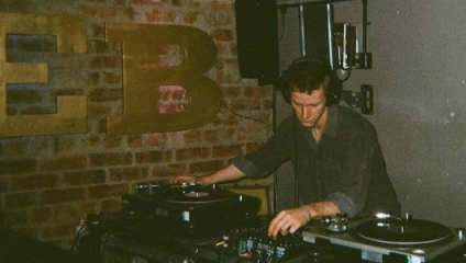 A DJ wearing headphones stands behind a set of turntables, adjusting the controls on a mixer. The background features a brick wall with large, partially visible letters in a metallic finish. The lighting is dim, creating a relaxed and intimate atmosphere.