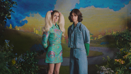 Two women standing in front of a painted backdrop featuring a whimsical cityscape. The woman on the left has long blonde hair with pink streaks and is wearing a colorful, patterned outfit. The woman on the right has dark hair, is wearing denim and has her hands behind her back.
