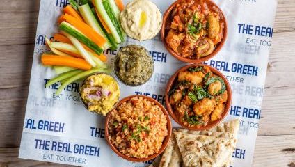 Veggie launch time platter. Lots of colourful baton veggies, 3 dipping ramekins, 3 larger bowls with Greek food and flat bread at the side