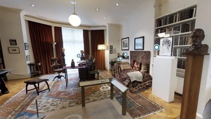 A cozy study room with a well-lit window adorned with long, dark curtains. The room features an ornate rug, a wooden desk with several items on it, a few chairs, bookshelves lining the wall, framed pictures, and a statuette on a stand.