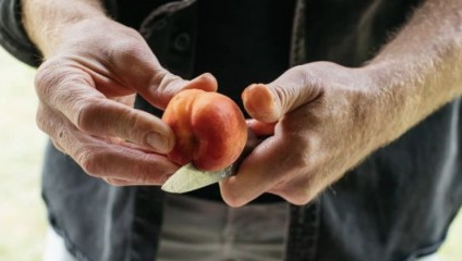Close-up of a person peeling a peach with a knife. The person is holding the peach in one hand and using a small knife to remove the skin. The background is blurred, focusing on the hands and the peach.