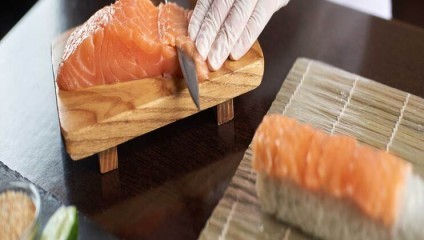 A person wearing a white glove uses a knife to slice raw salmon on a wooden board. In the foreground, a piece of salmon lies on top of rice on a bamboo mat. Various ingredients and tools for sushi preparation are visible on the dark surface.