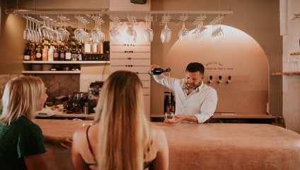 A bartender in a white shirt is pouring a drink into a glass for three female patrons sitting at a bar with hanging wine glasses above. The bar interior features a warm, cozy ambiance with shelves of bottles and taps visible in the background.