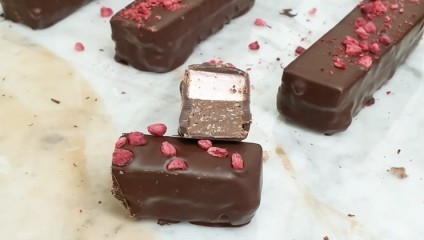 Several rectangular, chocolate-covered protein bars are displayed on a light-colored surface. One bar is cut in half, revealing a layered interior, while others are decorated with red, freeze-dried raspberries. Crumbs and chocolate pieces are scattered around.