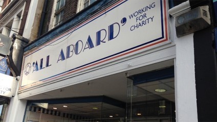 A storefront with a large sign above that reads ALL ABOARD WORKING FOR CHARITY in blue text. The facade is white with blue and red trim. The interior of the shop is partially visible through the glass windows and door.