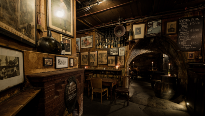 Dimly lit rustic bar with wooden furniture, vintage photographs on exposed brick walls, and candles creating a warm ambiance. A chalkboard menu lists Specials Reds on the right, and an archway leads to a cozy seating area with more framed photos on the wall.