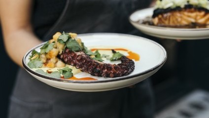 A person in a dark apron holds a plate with a grilled octopus tentacle garnished with herbs, potatoes, and a light sauce. Another dish, partially visible, is also being held, showing leafy greens and a browned surface. Both plates have elegant presentations.