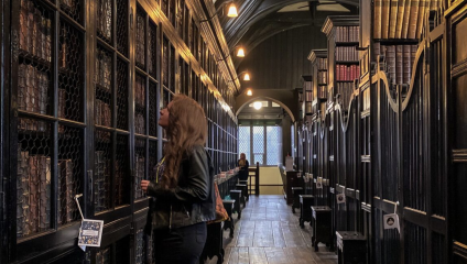 Inside the dark wood library loaded with books on shelves. Long haired lady standing in the foreground.
