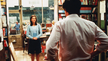 A woman in a pale blue sweater and dark skirt stands in a bookshop facing a man in a white shirt, who has his back to the camera. Shelves filled with books surround them, and a man wearing a blue shirt is seen through the window in the background.