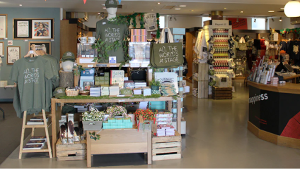 A bookstore display features a wooden table adorned with various books, hats, and gift items. A green hoodie and cap with matching text are showcased. Visible signs read “All the World’s a Stage.” In the background, more products and a board with posters can be seen.