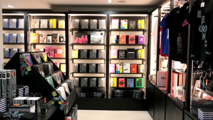 A retail store interior featuring neatly arranged shelves with colorful books and merchandise. T-shirts hang on the right, while the left displays racks of books and posters. The modern, well-lit space has a clean, organized layout with a variety of vibrant items.