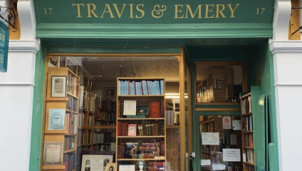 A bookstore named Travis & Emery with a green facade and gold lettering. The window displays various books and framed pictures. The interior, visible through the glass, is filled with shelves of books. The store is located at number 17.