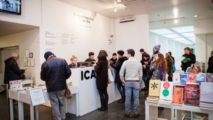 A group of people are gathered in the lobby of an art gallery, some standing in line while others browse books on display. A white reception desk with ICA on the front is staffed with two people assisting visitors. Various books and pamphlets are arranged on a nearby table.