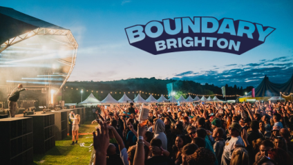 A vibrant crowd enjoys a live music performance at a festival. The stage is brightly lit with spotlights, and large white tents are set up in the background under a clear evening sky. The text Boundary Brighton is displayed prominently at the top of the image.