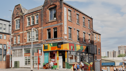 A vibrant, red-brick corner building with colorful murals on the lower facade featuring geometric patterns. It has large windows and a unique architectural style. People are sitting outside at tables under a small awning. Taller buildings and a cloudy sky are visible in the background.