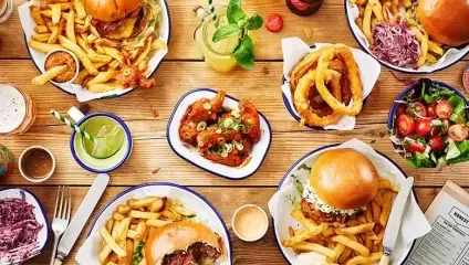 Lots of burgers, fries buckets, drinks and salads sitting on a wooden table top