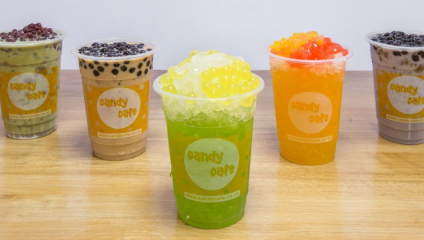 Five colorful cups of bubble tea are arranged on a wooden table. Each cup is labeled candy cafe and features a variety of toppings, including tapioca pearls and fruit jelly. The flavors appear to include matcha, chocolate, green apple, orange, and classic tea.