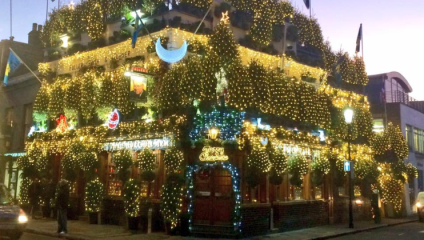 A building, likely a pub or restaurant, is adorned with extensive holiday lights and decorations. The lights cover most of the facade, creating a festive and vibrant atmosphere. People are seen near the entrance, and the surrounding street appears calm.